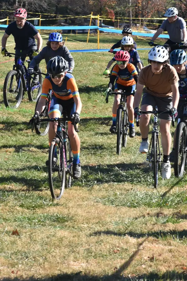 A wave of junior riders and parents rolling off the start line across open grass.