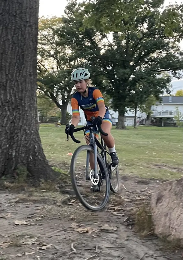 A young racer in an orange and blue kit railing a turn around a large oak at practice.